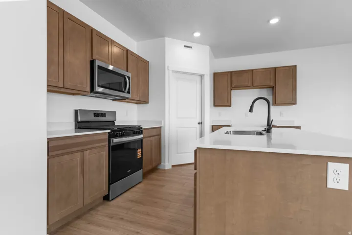 Kitchen featuring wood-finish cabinetry, light-toned countertops, stainless steel appliances, a center island with sink, and wood-finish flooring