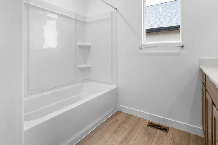 White bathtub with integrated shelving, wood-finish flooring, a single window, and a vanity with a light-toned countertop