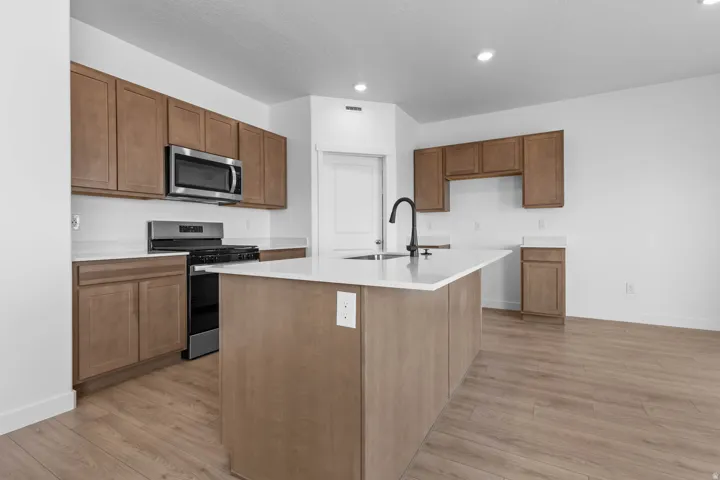 Modern kitchen featuring wood-finish flooring, white countertops, a central island with an integrated sink, stainless steel appliances, and wood cabinetry