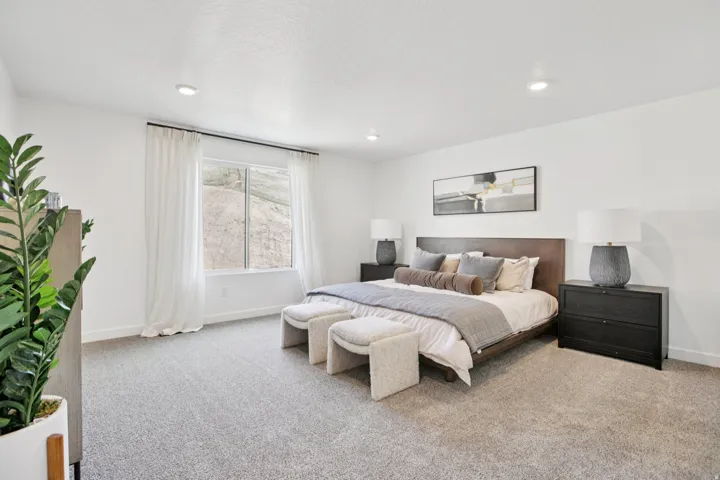 Recessed lighting illuminates a spacious room featuring a large window with white curtains and neutral-toned carpeting