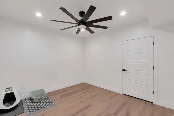 Recessed ceiling lighting, wood-finish flooring, white paneled door with black hardware, and a ceiling fan with integrated lighting