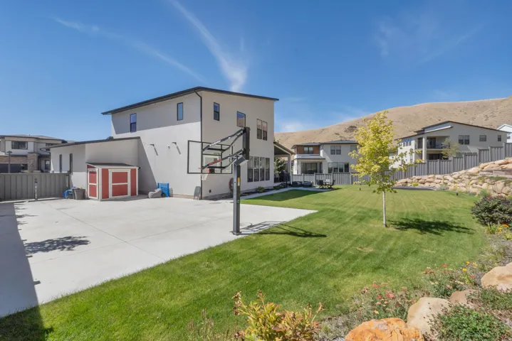 Expansive backyard featuring a large concrete patio, manicured lawn, and shed for additional storage and adjustable basketball standard.