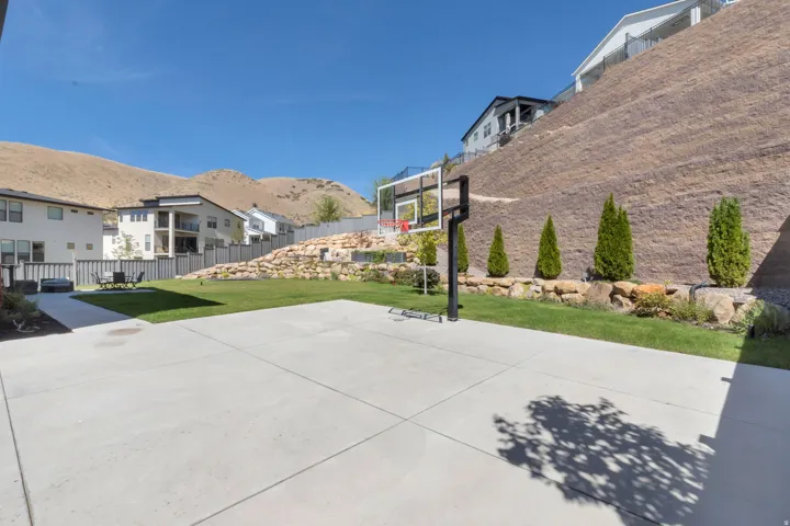 Expansive concrete patio with basketball hoop, manicured lawn, tiered landscaping with stone retaining walls, and mature evergreen plantings