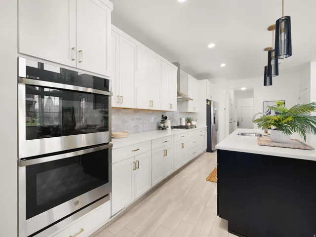 White shaker cabinetry with gold-finish hardware, stainless steel built-in double oven, light wood-finish flooring, and a large kitchen island with integrated sink