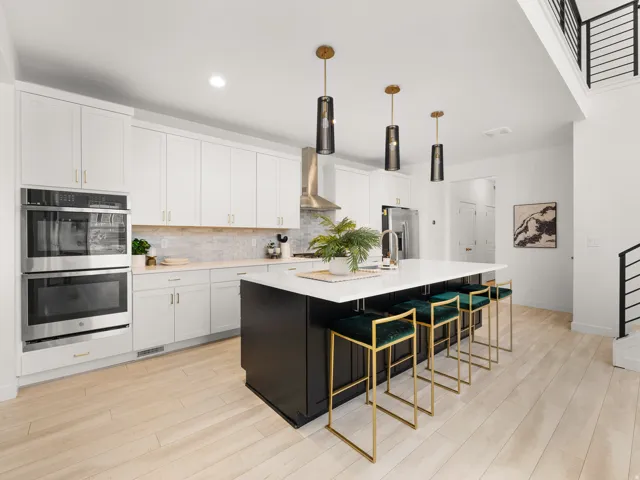 White cabinetry with gold-tone hardware, stainless steel double wall ovens, a large kitchen island with counter seating, wood-finish flooring, and a contemporary staircase with black railings