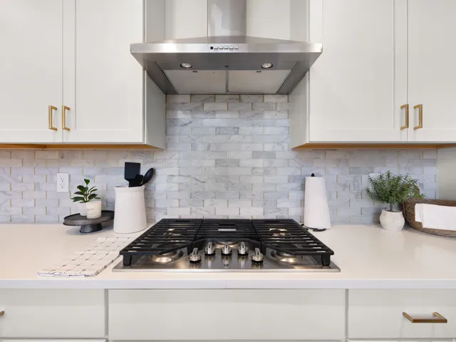 Stainless steel range hood, stone subway tile backsplash, gas cooktop, white shaker cabinetry, and light-toned countertops
