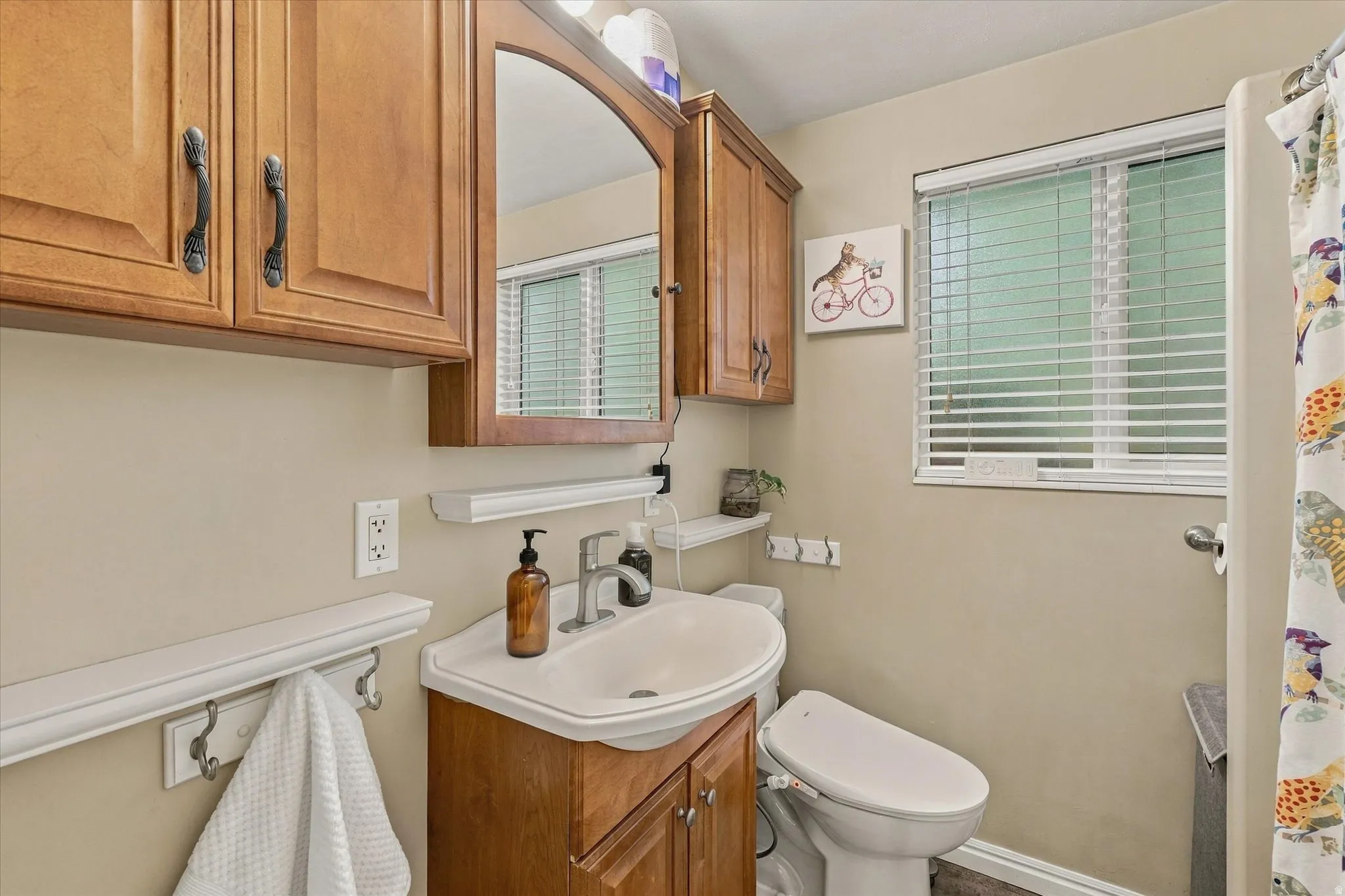 Bathroom featuring a wood-finish vanity with an integrated sink, coordinating upper cabinetry, an arched mirror, and a window with horizontal blinds