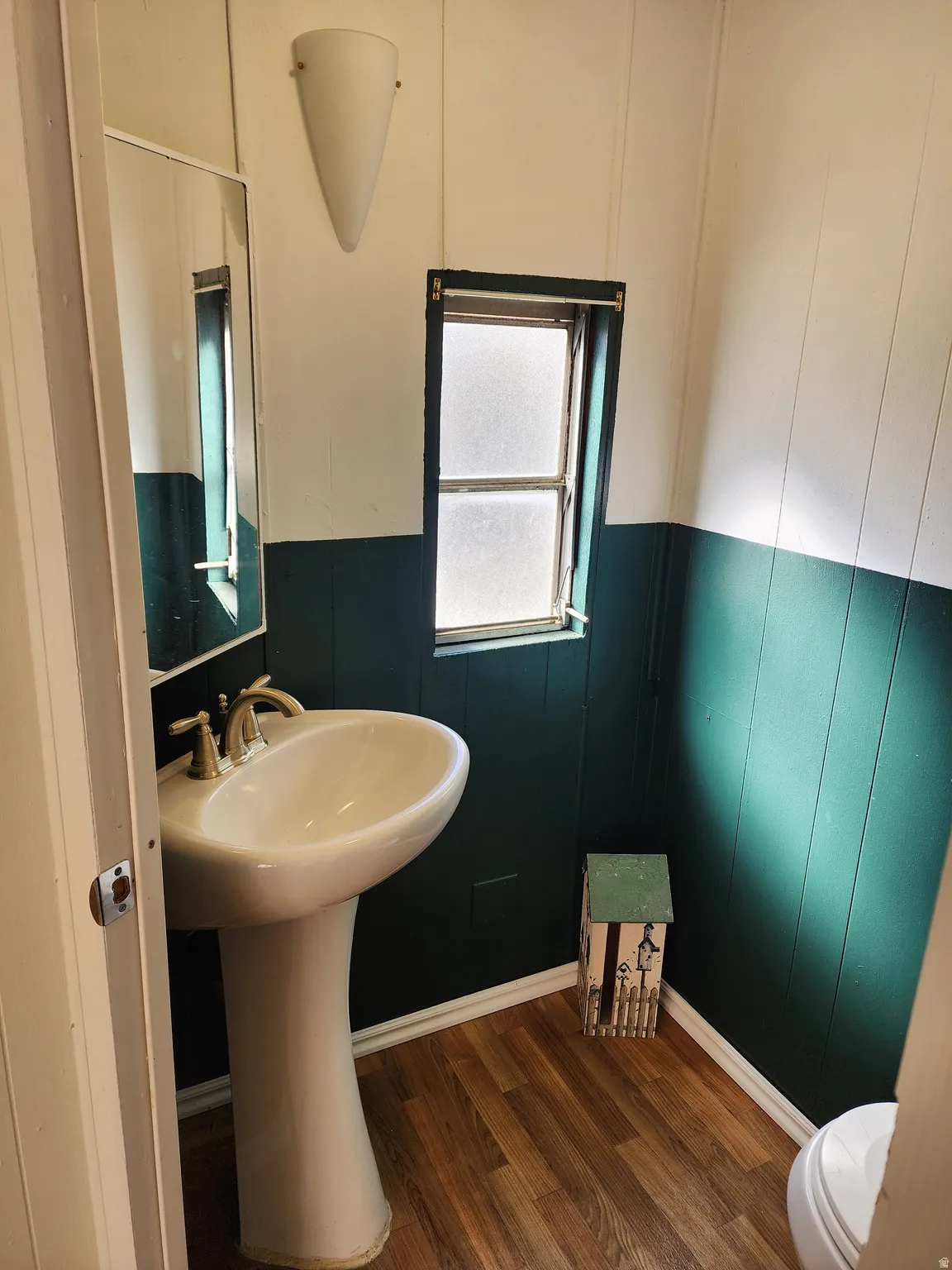 Pedestal sink with dual-handle faucet, wall-mounted mirror, wood-finish flooring, and a fixed window with privacy glass
