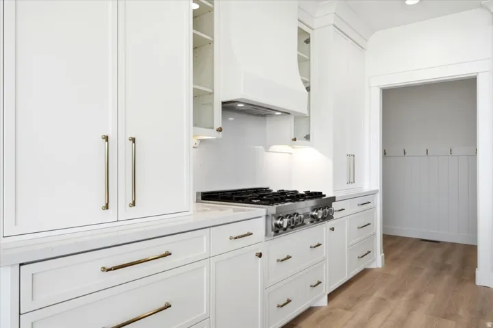 White shaker-style cabinetry with gold-tone hardware, professional-grade gas range, built-in range hood, light-toned wood-finish flooring, and white paneled walls with hooks