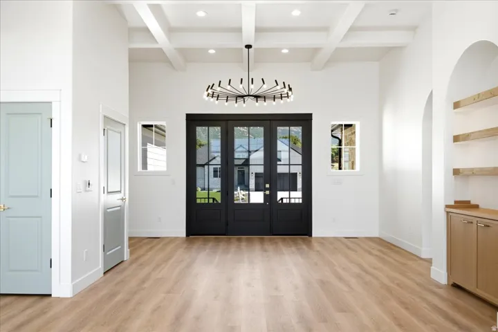 Double-entry doors with glass panels, wood-finish flooring, vaulted ceiling with exposed beams, built-in recessed shelving with integrated lighting, and a modern chandelier