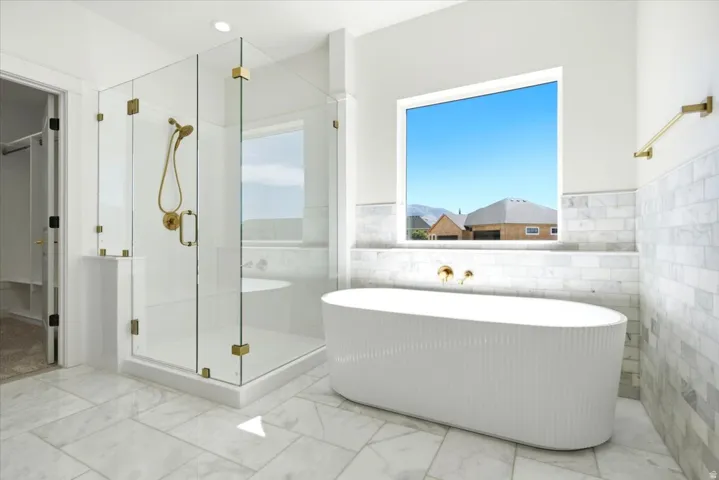 Primary ensuite bathroom. Freestanding soaking tub, walk-in shower with glass enclosure, brass fixtures, marble-look tile flooring, and half-tiled walls