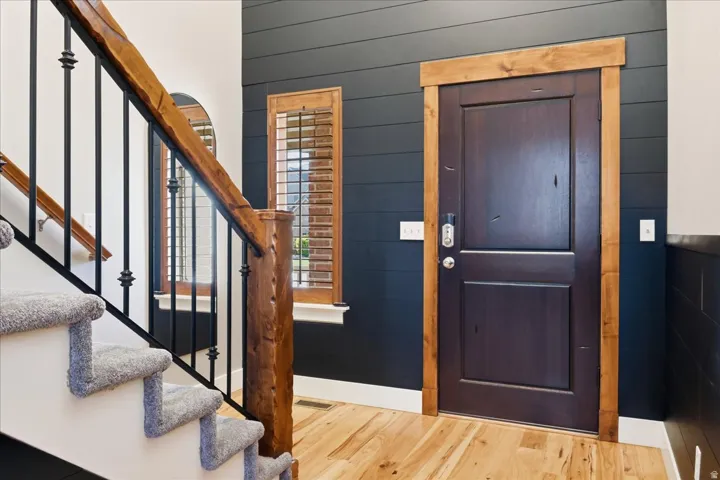 Hardwood flooring, dark wood-paneled wall, dark wood door with light wood trim, light wood staircase railing with black metal spindles, and a window with wood shutters
