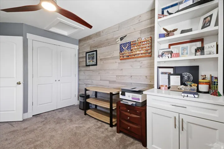 Built-in shelving, white cabinetry with silver-tone hardware, wood-finish accent wall, ceiling fan with integrated lighting, and neutral carpeting