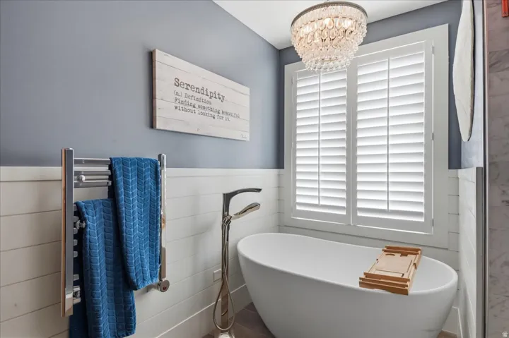 Freestanding soaking tub with floor-mounted faucet, plantation shutters, crystal chandelier, and shiplap wall accent