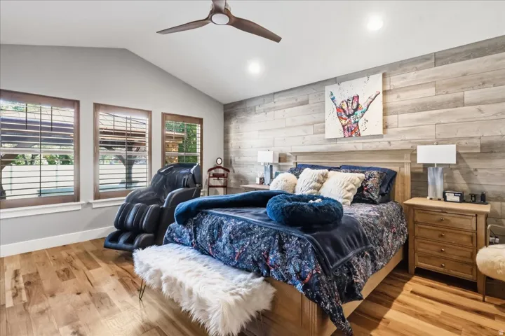 Vaulted ceiling with a ceiling fan, three windows with wood blinds, wood-finish flooring, and a wood plank accent wall