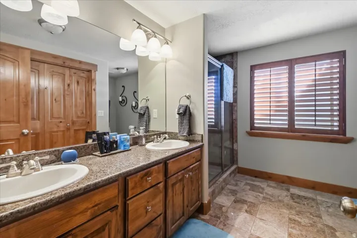 Dual vanity with integrated sinks and granite-look countertops, wood-finish cabinetry, tile flooring, glass-enclosed shower, and wood-framed window with plantation shutters