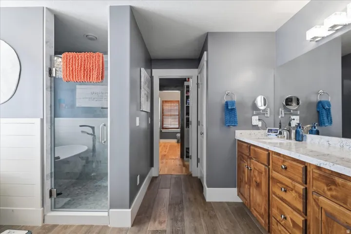 Glass-enclosed shower with a freestanding tub, dual vanity with wood cabinetry, stone-finish countertop, and wood-finish flooring