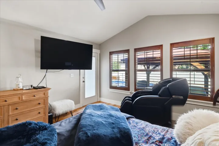 Vaulted ceiling with a ceiling fan, three windows with wood frames, wood-finish flooring, and a glass-paneled door to patio from primary bedroom