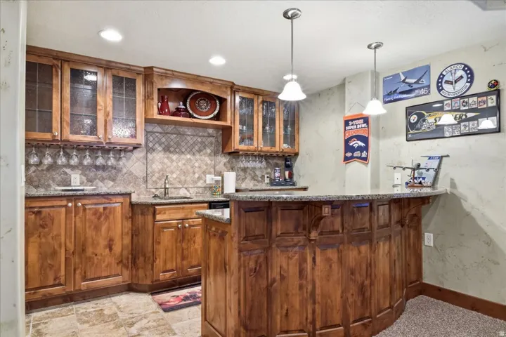 Custom wood cabinetry with glass-front uppers, stone countertops, stone tile backsplash, recessed lighting, and pendant lighting