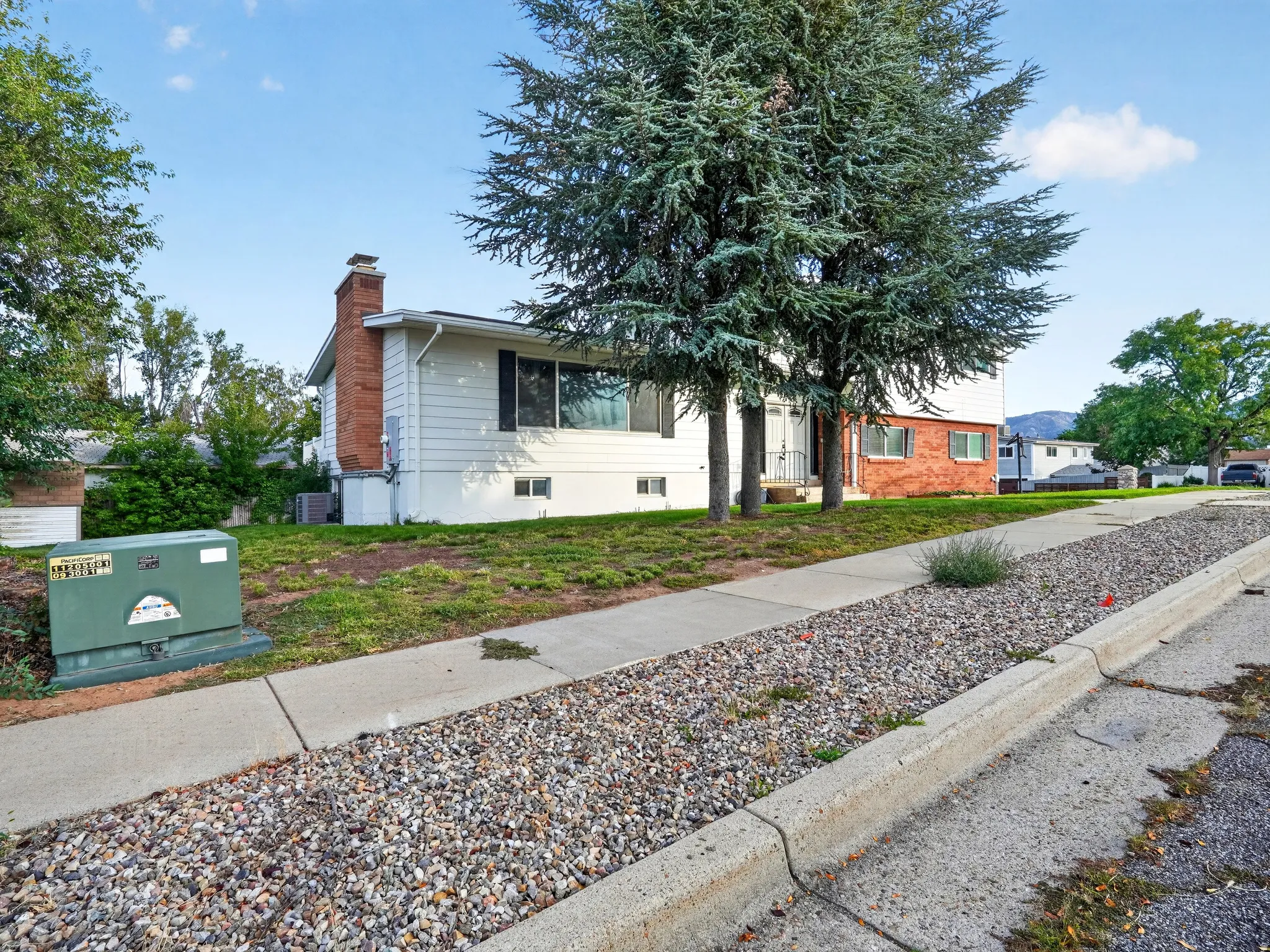White siding exterior with brick accents, mature trees, and a concrete sidewalk