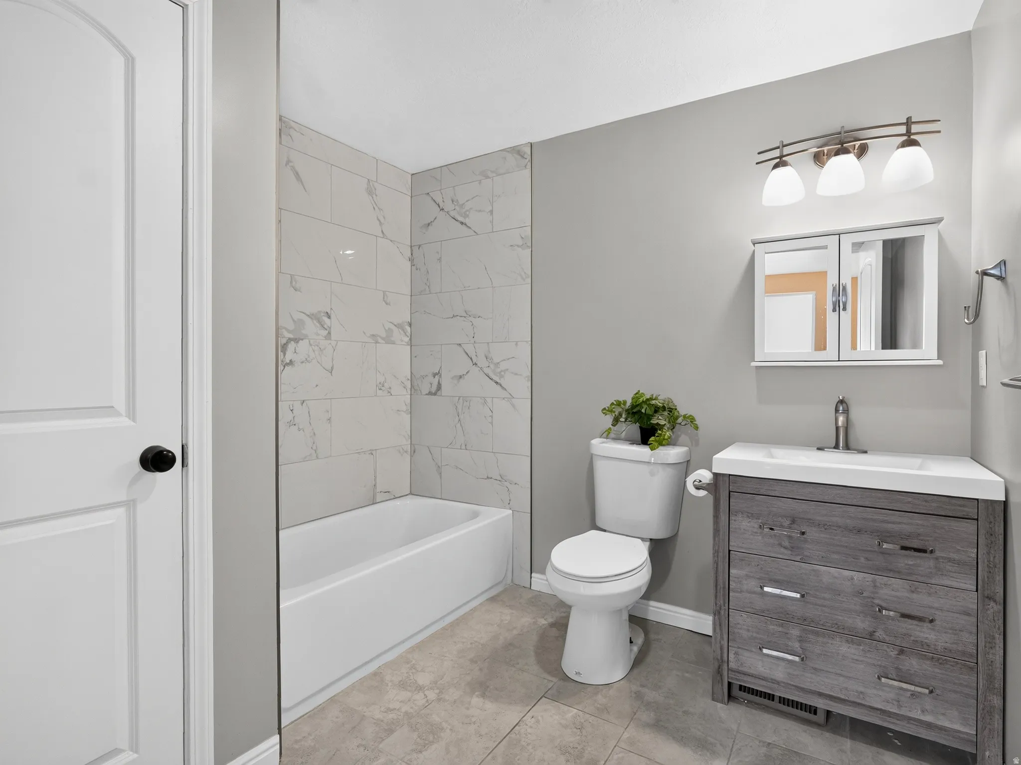 Bathroom featuring a white tub with marble-look tile surround, a distressed wood-finish vanity with an integrated white sink, a modern toilet, and a medicine cabinet with mirrored doors