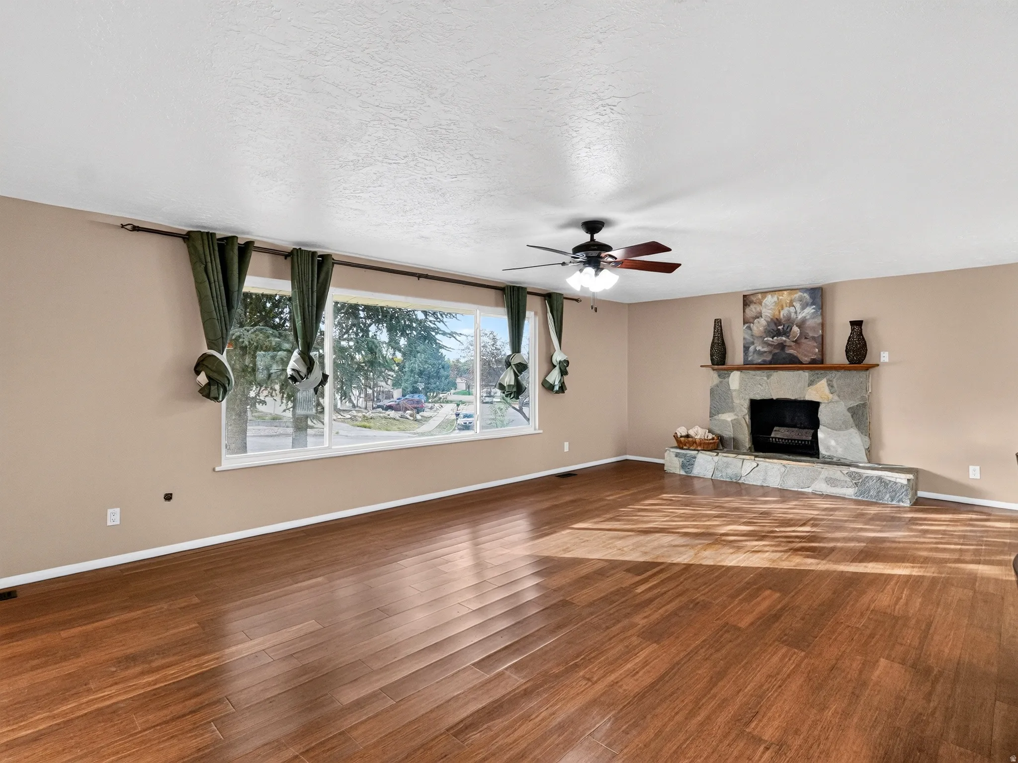 Wood-finish flooring, stone-clad fireplace, large picture window, and ceiling fan with lighting