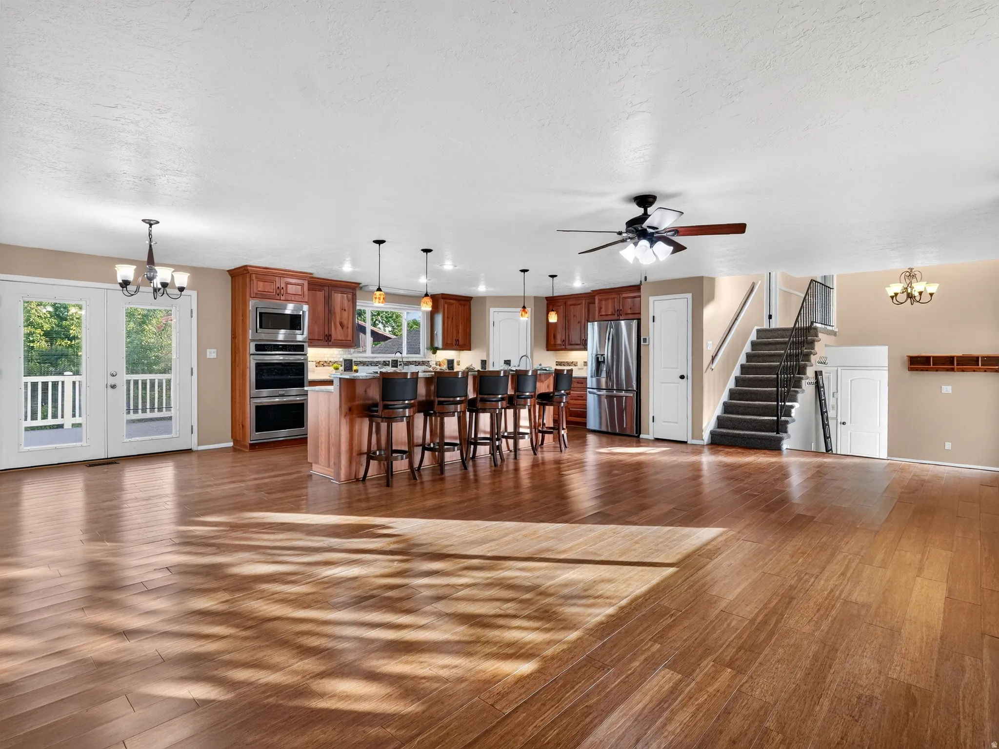 Open-concept living space featuring wood-finish flooring, a kitchen island with seating, stainless steel appliances, and a staircase with dark carpeting