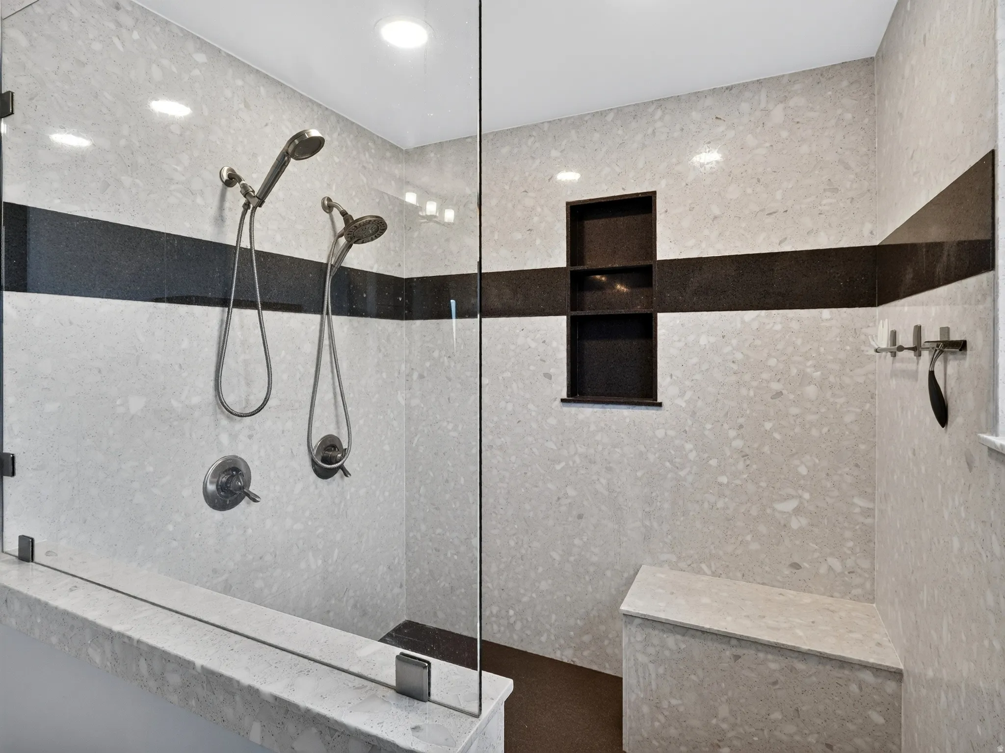 Spacious shower with stone-finish tile, dual shower heads, built-in shelving, glass enclosure, and integrated bench seating