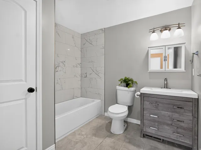 Bathroom featuring a white tub with marble-look tile surround, a distressed wood-finish vanity with an integrated white sink, a modern toilet, and a medicine cabinet with mirrored doors