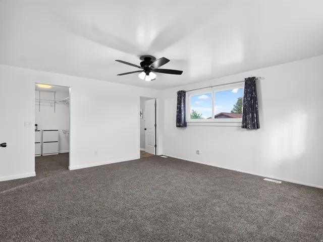 Spacious room featuring carpeting, a ceiling fan with integrated lighting, a large window, and an open doorway to a walk-in closet with built-in shelving