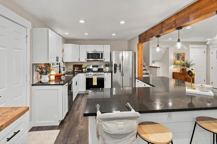 Kitchen featuring stainless steel appliances, dark stone countertops, white shaker cabinetry, wood-finish flooring, and exposed wood beam accents