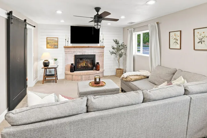 Brick-faced fireplace with a hearth, ceiling fan with integrated lighting, recessed lighting, white shiplap accent wall, and a dark barn door