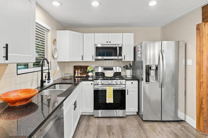 Kitchen featuring white shaker cabinetry, stainless steel appliances, dark countertops, a tile backsplash, and wood-finish flooring