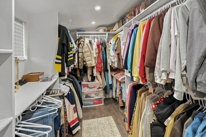 Custom closet system with white shelving, integrated hanging rods, recessed lighting, wood-finish flooring, and a window for natural illumination