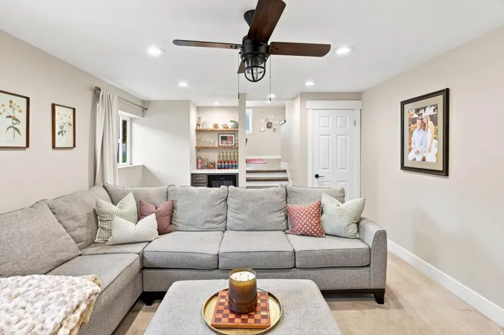 Recessed lighting, wood-finish ceiling fan with caged light fixture, built-in wet bar with wood shelving, compact refrigerator, and staircase with white risers