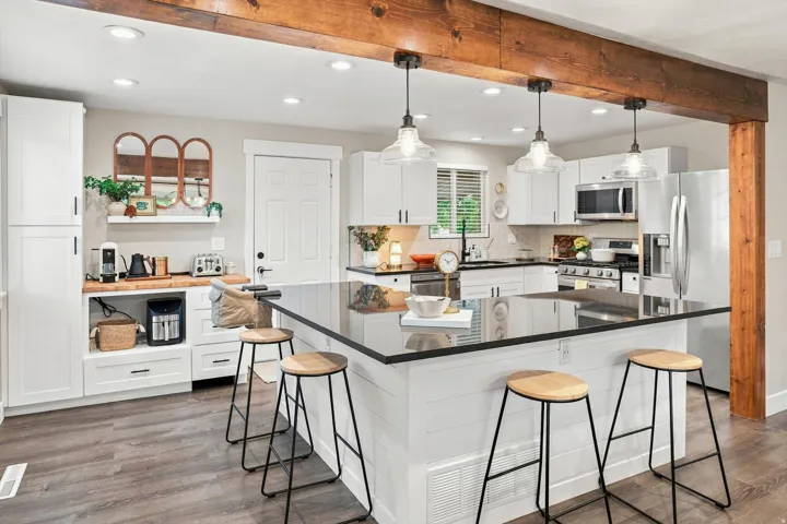 Open-concept kitchen featuring wood-finish flooring, white cabinetry, a large kitchen island with a dark countertop, stainless steel appliances, and exposed wood beams