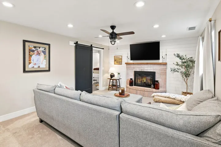 Corner fireplace with brick surround and wood mantle, shiplap accent wall, ceiling fan, recessed lighting, and barn door