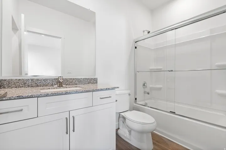 Bathroom featuring a white vanity, stone-finish countertop, single basin sink, and a shower-tub combination with glass enclosure