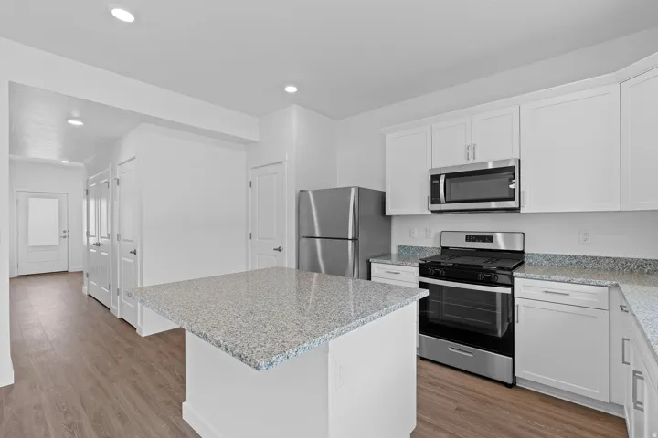 Modern kitchen featuring white cabinetry, stainless steel appliances including a gas range and microwave, speckled quartz countertops, a center island with breakfast bar seating, and wood-finish flooring throughout the open living space
