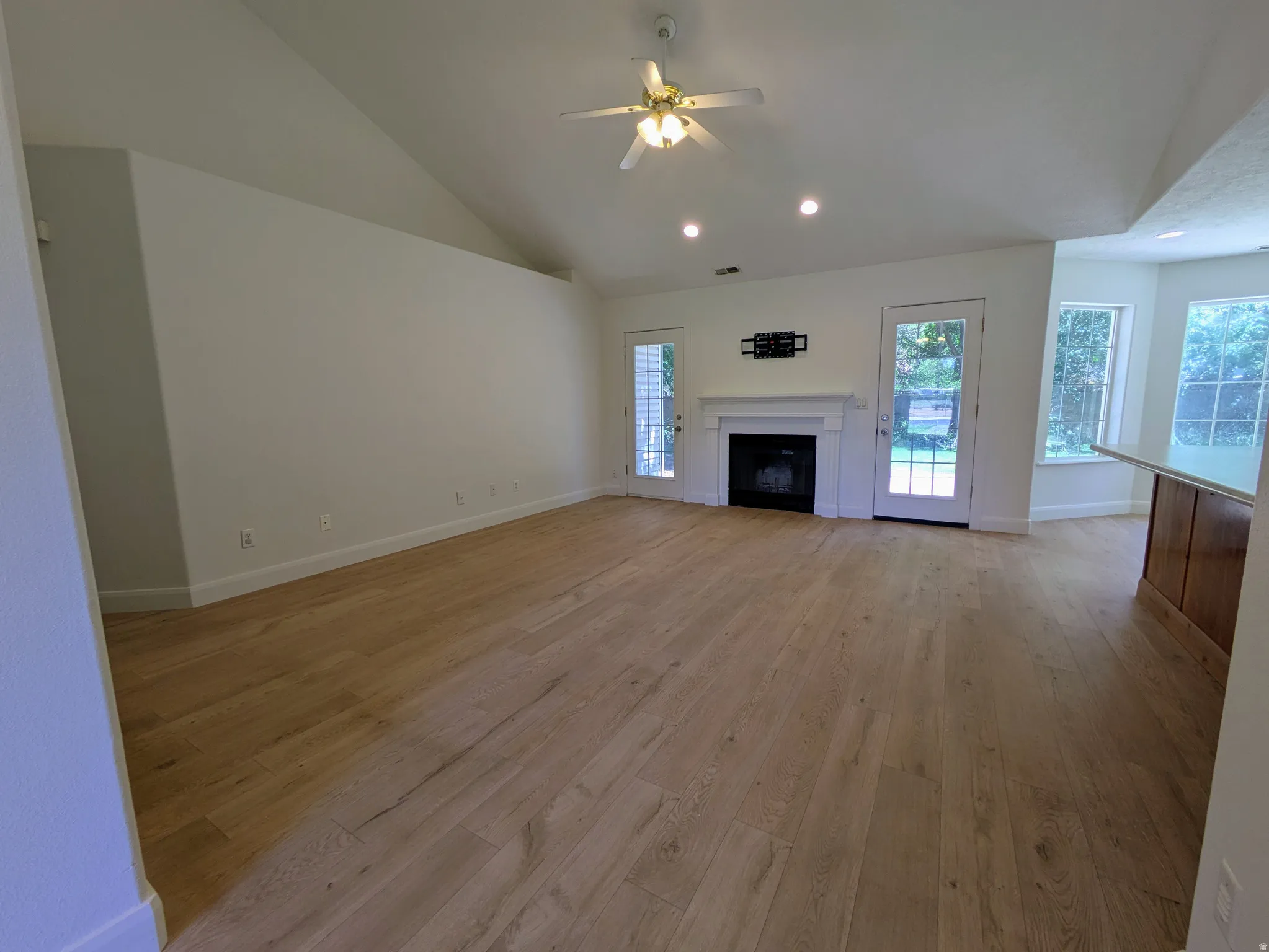 Vaulted ceiling with a ceiling fan, wood-finish flooring, fireplace with a mantel, two glass-paneled doors, and built-in breakfast bar