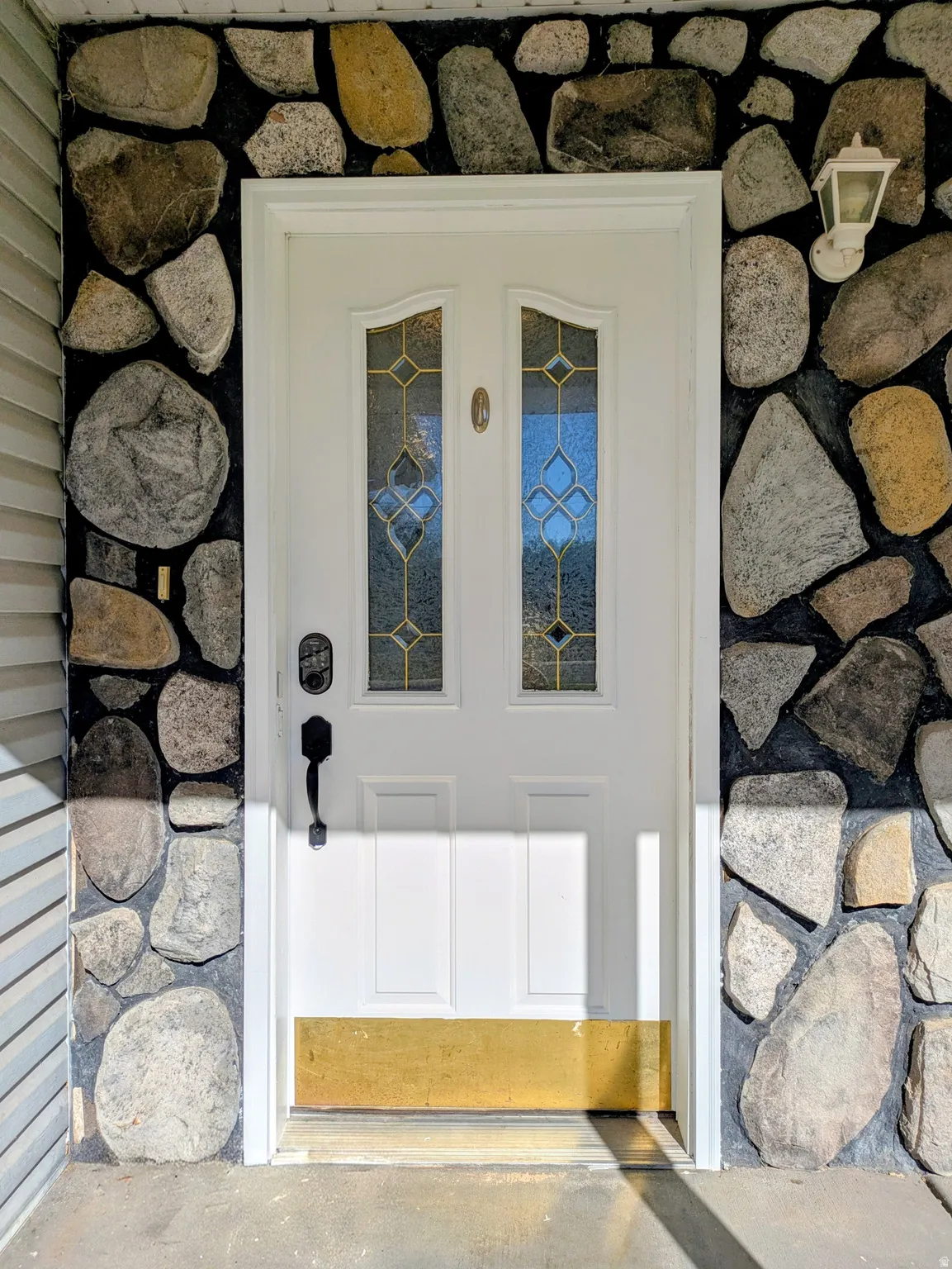 White entry door with decorative glass inserts, surrounded by a stone facade and featuring a wall-mounted exterior light fixture