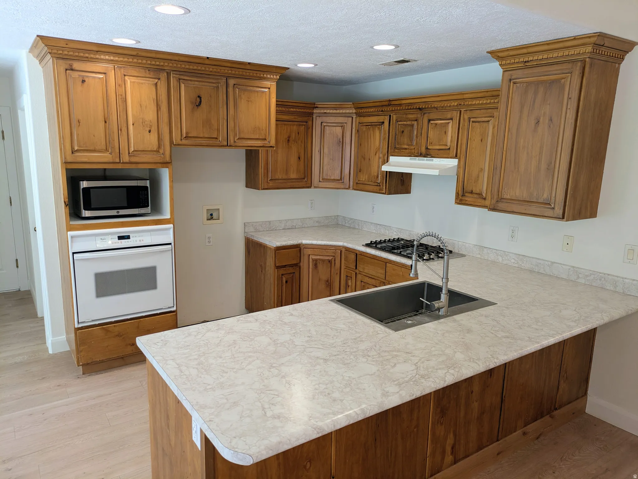 Wood-finish cabinetry with crown molding, integrated built-in oven, gas cooktop, and a stainless steel sink with a high-arc faucet