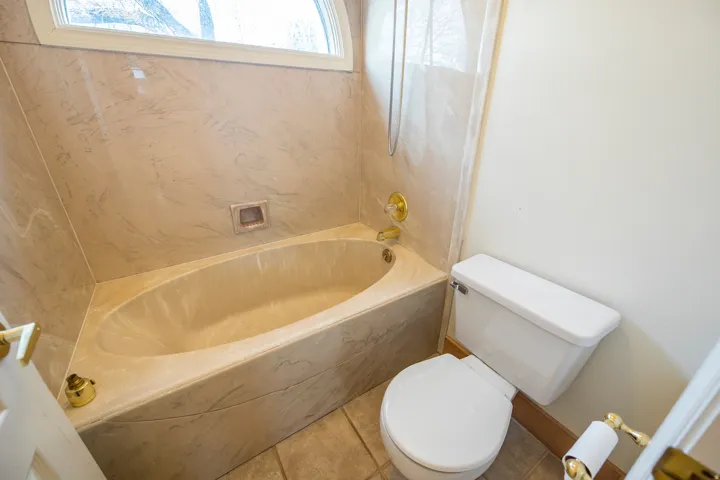 Built-in soaking tub with a tiled surround, gold-tone fixtures, and a curved window above
