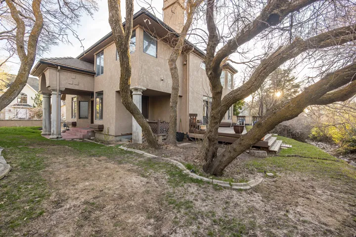 Stucco exterior with a tiled roof, featuring a covered patio with stately columns, a multi-level wood deck, and mature trees