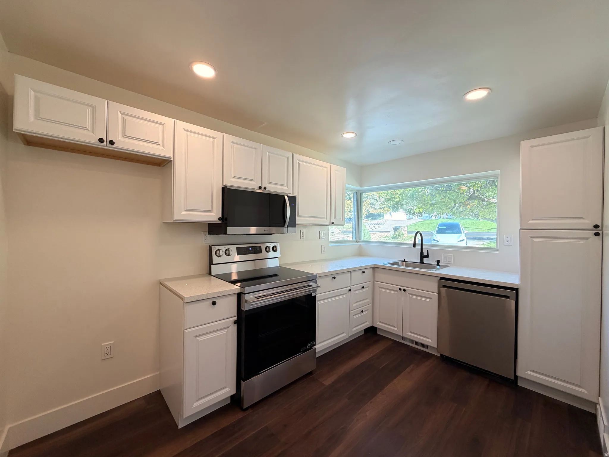 White shaker cabinetry, dark wood-finish flooring, stainless steel appliances, recessed lighting, and a large picture window