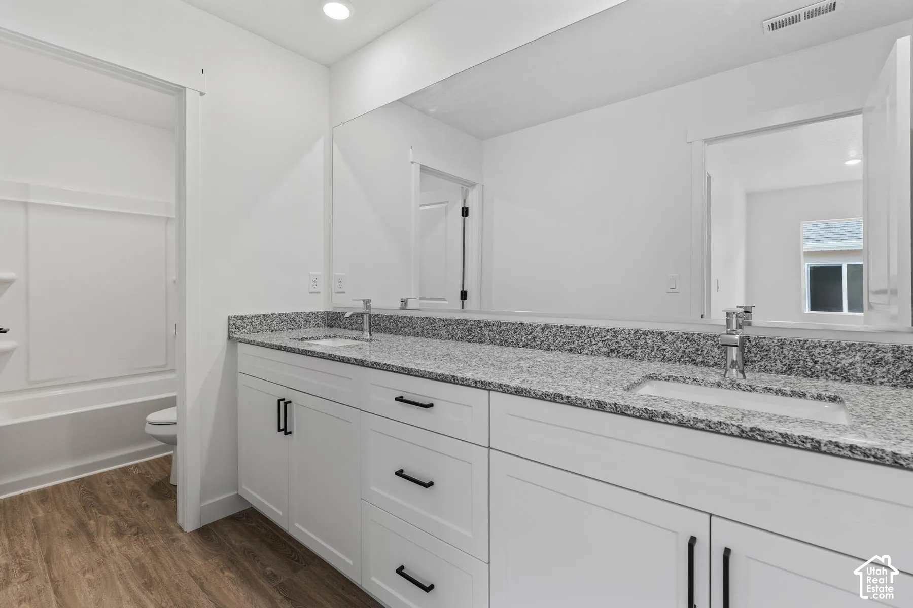 Dual undermount sinks with chrome fixtures, granite countertop, white shaker-style cabinetry with matte black hardware, integrated bathtub/shower combination, and wood-finish flooring