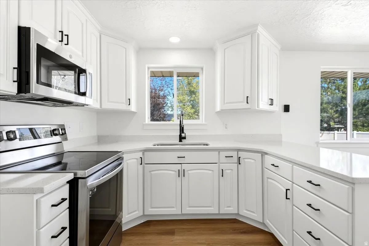 White cabinetry with black hardware, stainless steel appliances, light-toned countertops, wood-finish flooring, and a single basin sink with a black faucet