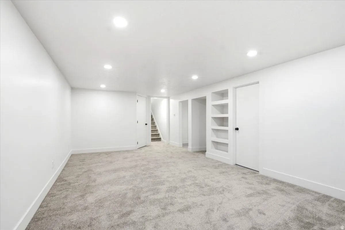 Spacious room featuring recessed lighting, light-toned carpet, white walls, built-in shelving, and a doorway opening to a staircase