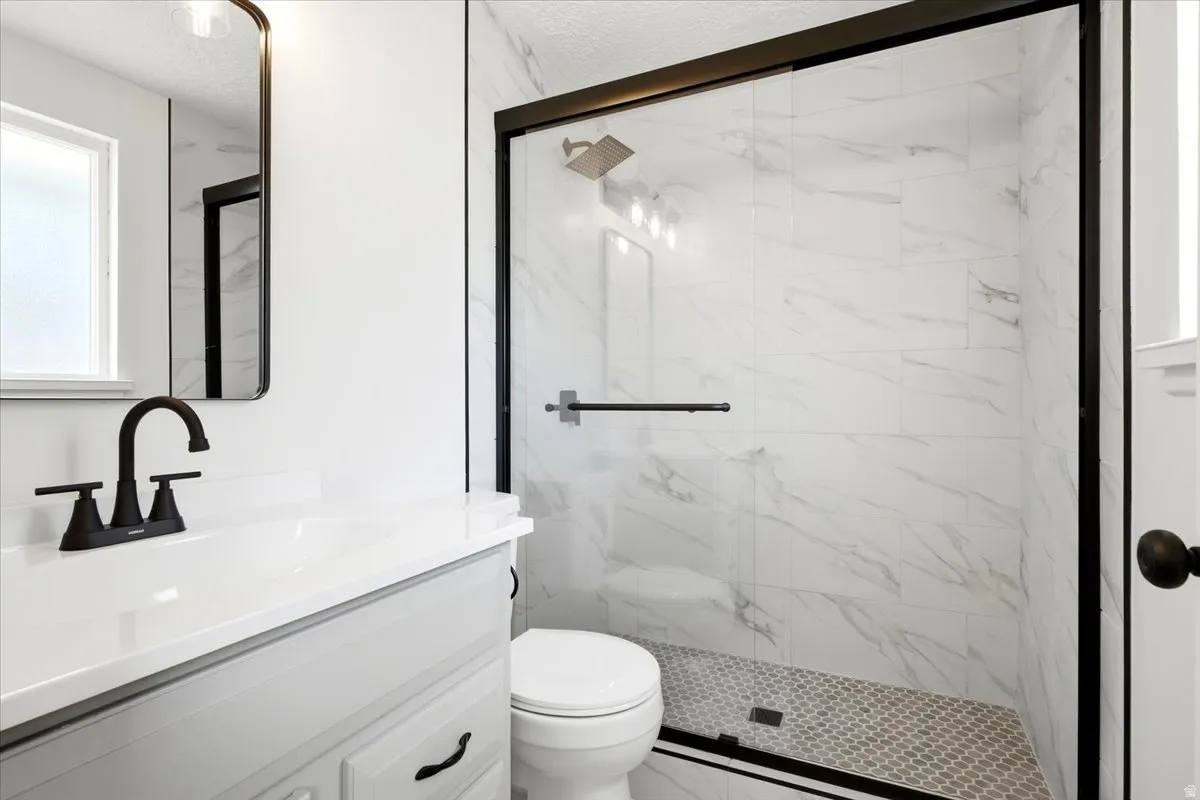 Bathroom featuring a single vanity with a white countertop, matte black fixtures, a framed mirror, and a glass-enclosed shower with tiled walls and a mosaic-tiled floor