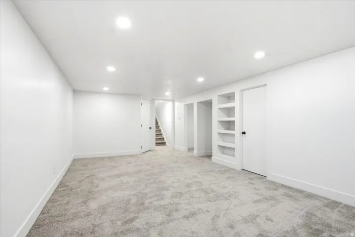 Spacious room featuring recessed lighting, light-toned carpet, white walls, built-in shelving, and a doorway opening to a staircase